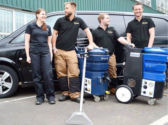 This is a photo of Abbots Langley Carpet Cleaning carpet cleaners (three men and one woman) standing in fromt of their black van, with two steam cleaning carpet machines next to them